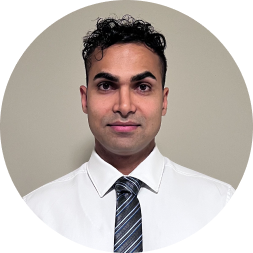 Professional headshot of a man with dark hair wearing a white dress shirt and striped navy tie against a neutral beige background.