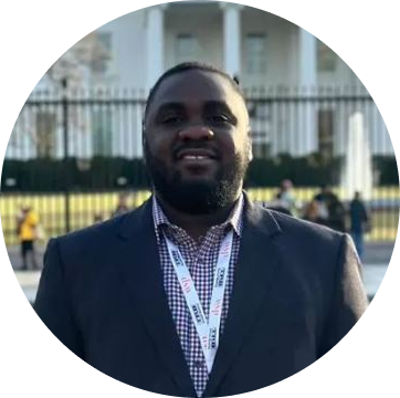 Professional headshot of a man wearing a dark blazer and blue checkered shirt with a conference badge, photographed outdoors with a building visible in the background.
