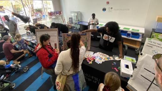 University researchers demonstrate interactive STEM projects to visitors at an indoor outreach event, with children engaging with educational games and displays on tables.