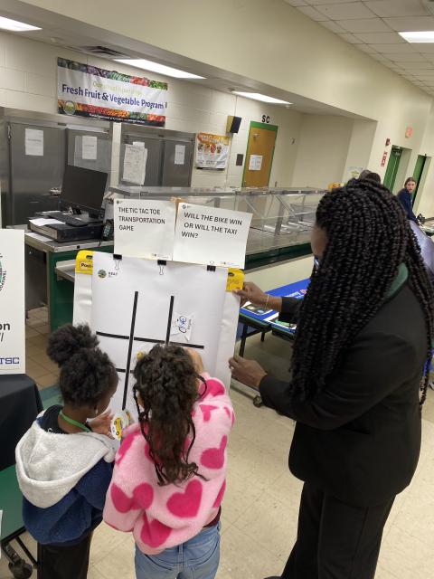 Two children play tic-tac-toe at a game station while an adult supervises in a school cafeteria during an educational program.