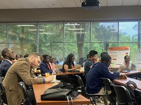 A diverse group of researchers and students seated at tables during a seminar or workshop in a classroom with large windows overlooking trees. Research posters are visible on the wall.