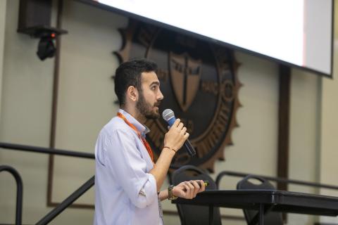 Man in light blue shirt speaking at podium with microphone in university lecture hall setting.