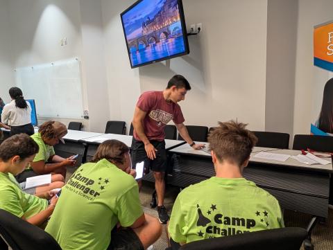 An instructor in a maroon shirt leads a discussion with several students wearing green Camp Challenger shirts seated around a table in a classroom setting.