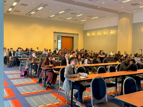 A conference room filled with attendees seated at tables during what appears to be an academic or professional event, with yellow walls and colorful patterned carpeting.