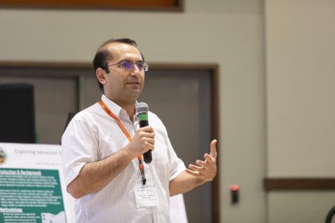 Man wearing glasses and orange lanyard speaking at podium with microphone during academic presentation.