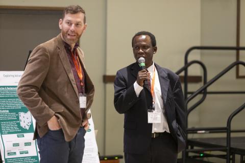 Two researchers present at a conference, one holding a microphone while speaking and the other standing beside a research poster displaying data and findings.
