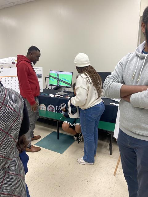 Students gather around a table examining a computer monitor displaying a map interface during what appears to be a university research or technology demonstration event.