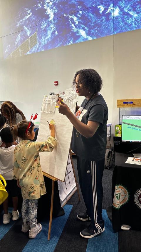 An educator demonstrates a drawing or diagram to young students in a classroom while satellite imagery displays on a screen above.