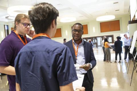 Researchers wearing lanyards engage in conversation at a university conference or research event in a modern building hallway.