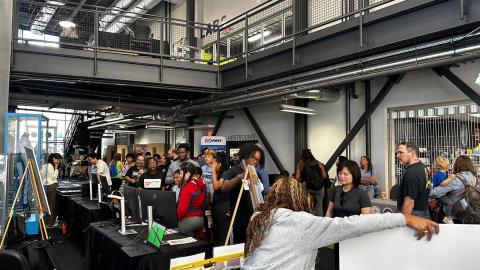Large group of people gathered in an industrial workshop space with exposed beams and metal railings, examining robotics equipment and computer stations at what appears to be a FIRST Robotics event or competition.