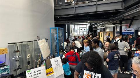 Crowded indoor event at a research facility with visitors viewing displays, including glass cases with artifacts and a drawing activity station.