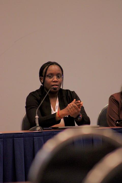 A woman wearing glasses and a black blazer speaks at a panel table with a microphone, wearing a conference badge and colorful wristband.