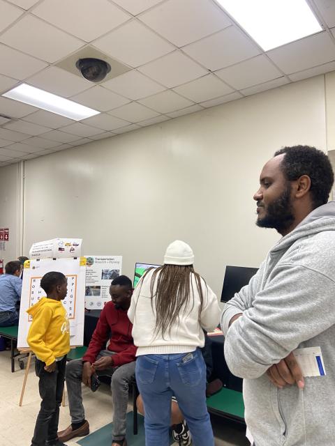 Group of students and adults engaged in discussion around a presentation poster in a classroom setting with fluorescent ceiling lights.
