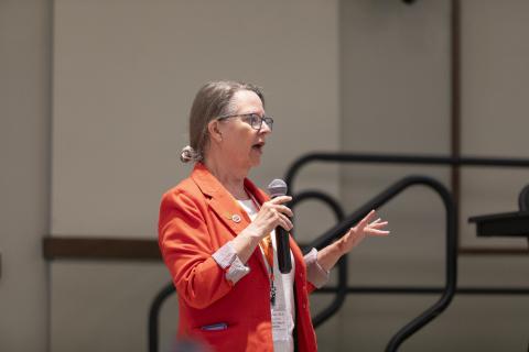 Woman in red blazer speaking at a microphone during a university presentation, wearing glasses and a name badge.