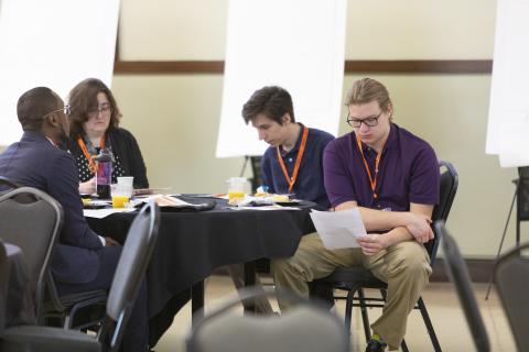 Four people seated at a black conference table during what appears to be a panel discussion or meeting, with orange lanyards visible and beverages on the table.