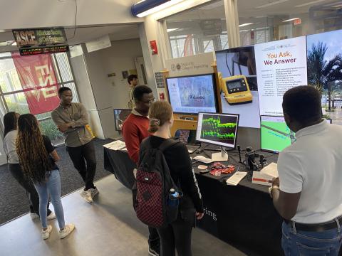 Students gather around a display table in a university engineering lab featuring monitors, laptops, and demonstration equipment during an open house event.