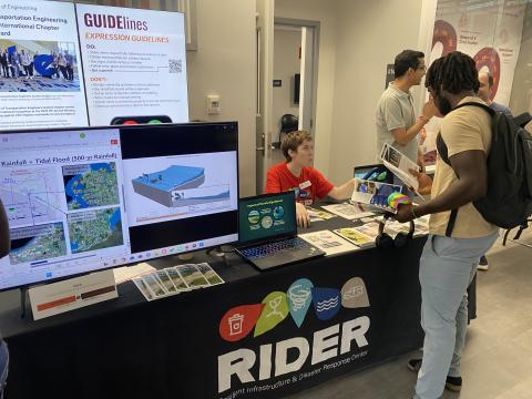 Students engage with researchers at a RIDER (Resilient Infrastructure & Disaster Response Center) booth displaying presentation materials on rainfall, tidal flooding, and infrastructure design.