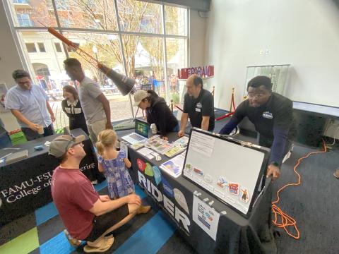 University staff and visitors engage with an interactive robotics display at an indoor event, with large windows overlooking a campus plaza in the background.