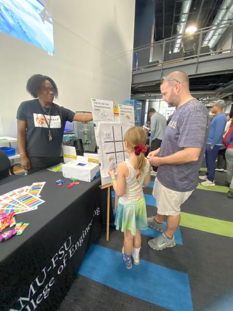 A presenter at a university engineering booth demonstrates an interactive tic-tac-toe transportation game to a young child and adult visitor at an indoor event.