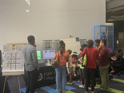 Visitors gather around a display table at a FAMU-FSU College of Engineering event, examining a tic-tac-toe transportation game and computer demonstration.