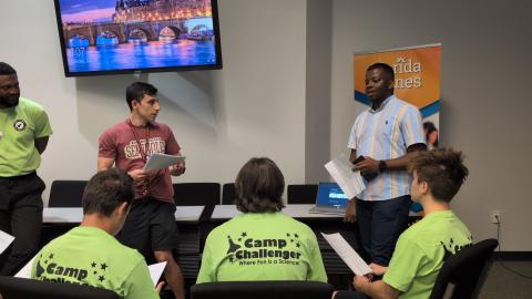 Presenter in striped shirt addresses Camp Challenger participants wearing bright green shirts in a classroom with a bridge scene displayed on the wall monitor.