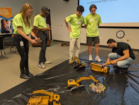Students in bright yellow shirts demonstrate robotic excavators and machinery on a black mat in a classroom, with one person crouching to adjust equipment while others observe.