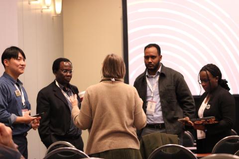 Five professionals wearing conference badges engage in a discussion during a networking event in front of a curved pink and white graphic backdrop.