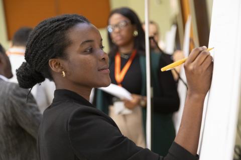 Woman in black shirt writing on whiteboard with pencil while others observe in background at indoor event.