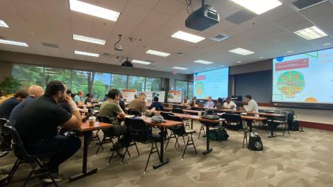 Academic conference or seminar in a university classroom with attendees seated at tables facing a projection screen displaying presentation slides.