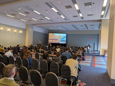 Conference room filled with attendees seated in rows facing a projection screen displaying a presentation slide with a building image and "Thank You" text.