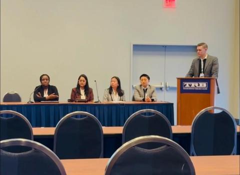A man speaks at a wooden podium while four panelists sit at a table with a blue skirt and microphones during an academic panel discussion.