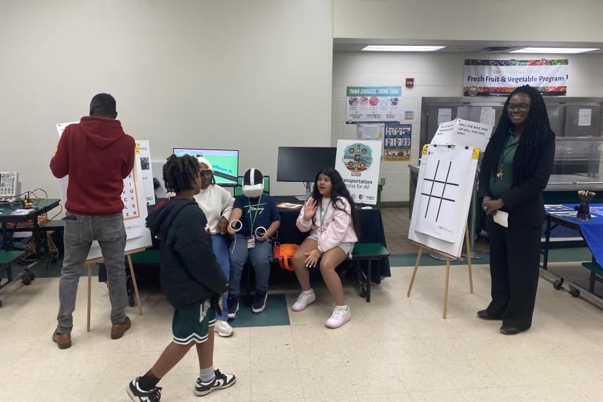 Students and an adult participate in an interactive educational activity in a classroom, with one student gesturing while seated and others standing nearby observing a presentation board.