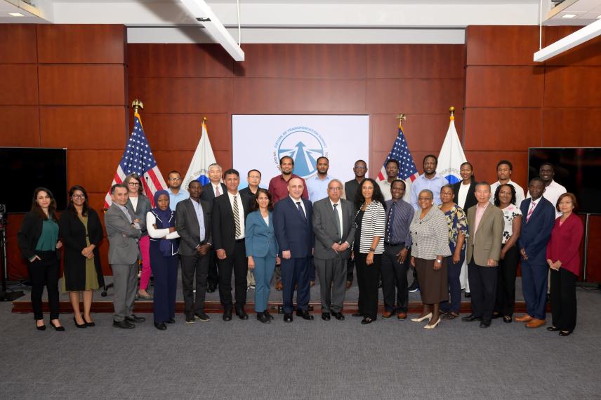 Group portrait of approximately 30 professionals in business attire standing in front of a red wood-paneled wall with American flags and an official seal displayed behind them.