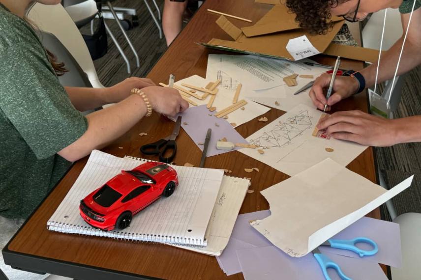 Students gathered around a wooden table work on an engineering design project with sketches, wooden materials, scissors, and a red toy car model.