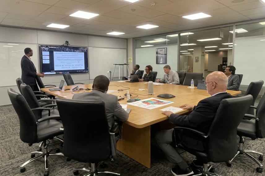 Business professionals in a conference room seated around a wooden table while one man presents at a wall-mounted display screen.