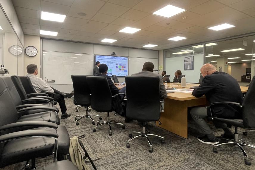 University research meeting in progress with six participants seated around a conference table, one person presenting on a wall-mounted screen.