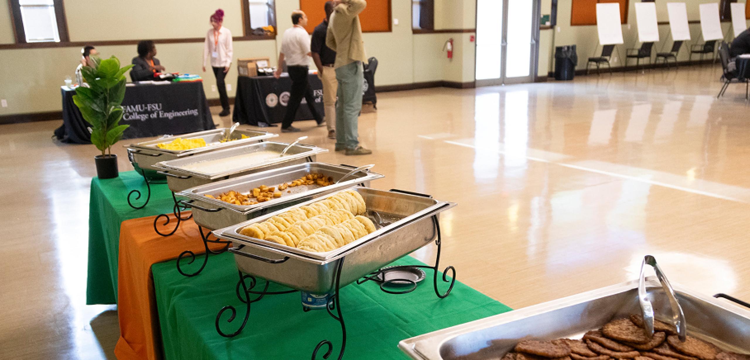 View of a large room with buffet trays in front. There's people sitting at tables and walking around further into the room.