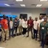 Nine people wearing lanyards stand in a classroom with computer equipment and displays, posing for a group photo.