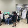 Students and staff participate in an interactive educational activity in a school cafeteria, with game boards and informational posters displayed on easels.