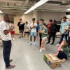 An instructor leads a group of diverse young people gathered in a laboratory or workshop space, demonstrating or discussing materials near a cardboard box on the floor.