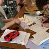 Students gathered around a wooden table work on an engineering design project with sketches, wooden materials, scissors, and a red toy car model.