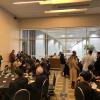 Networking event in a modern conference room with attendees seated at tables and standing, with beverages and refreshments visible.