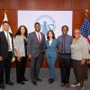 Six professionals stand in front of a "Future of Transportation Summit" banner, flanked by American flags and state flags in a formal setting.