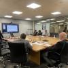 Business professionals in a conference room seated around a wooden table while one man presents at a wall-mounted display screen.